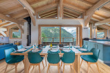 Salle à manger avec sa grande table, chaises bleues, plafond cathédral, poutres apparentes et grandes baies vitrées, vue sur les montagnes
