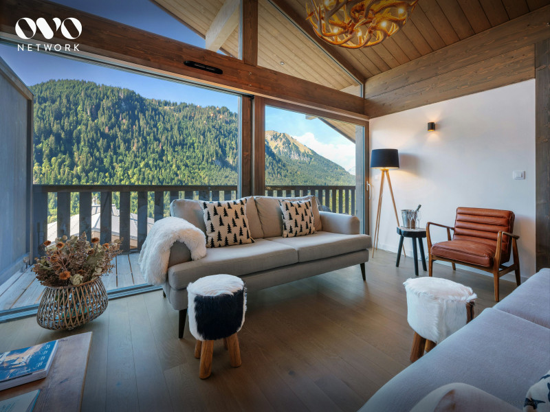 Salon très lumineux avec canapé et fauteuil, grande baie vitrée donnant accès au balcon et vue sur les montagnes